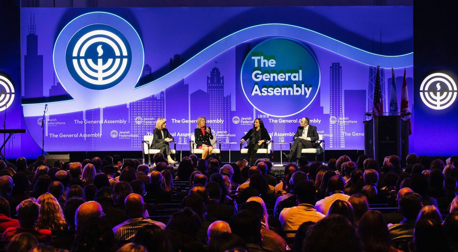 a large group of people are sitting in front of a stage at the JFNA general assembly.