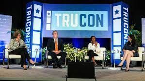 a group of people are sitting on a stage in front of large trucon logo.