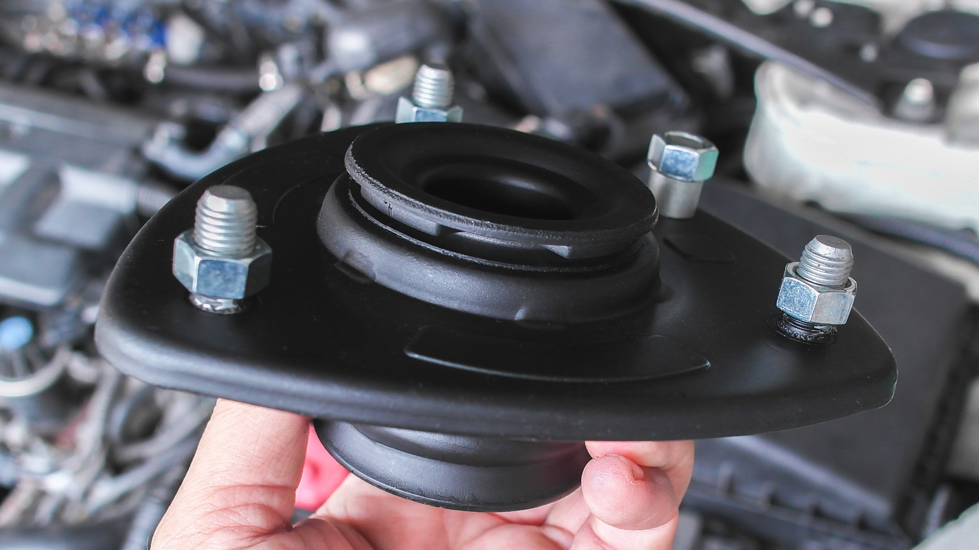 Black car strut mount with rubber bushing and bolts, held by a person's hand with engine in the background.
