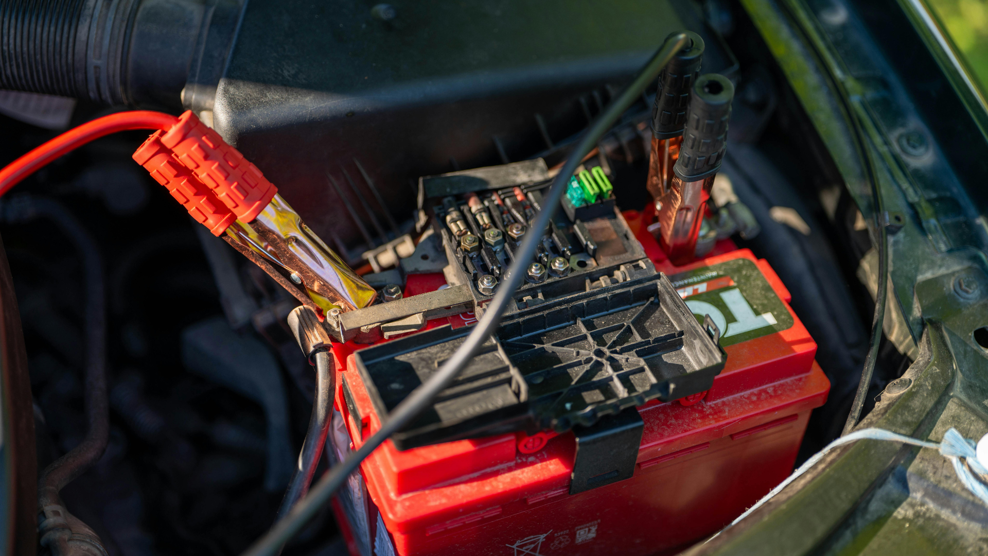 Red car battery with jumper cables attached, in an open engine compartment.