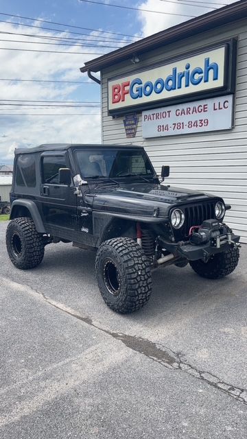Black Jeep Wrangler parked in front of a BFGoodrich garage. Large tires and winch visible.