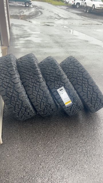 Four new black all-terrain tires leaning on a wet paved surface outdoors.