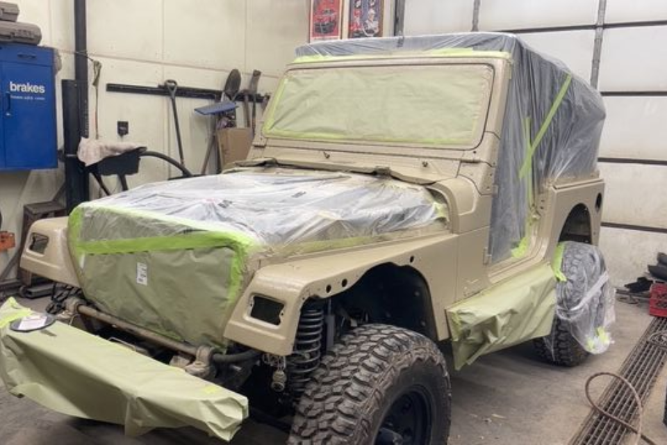Tan vehicle body in a garage, taped and covered for painting.
