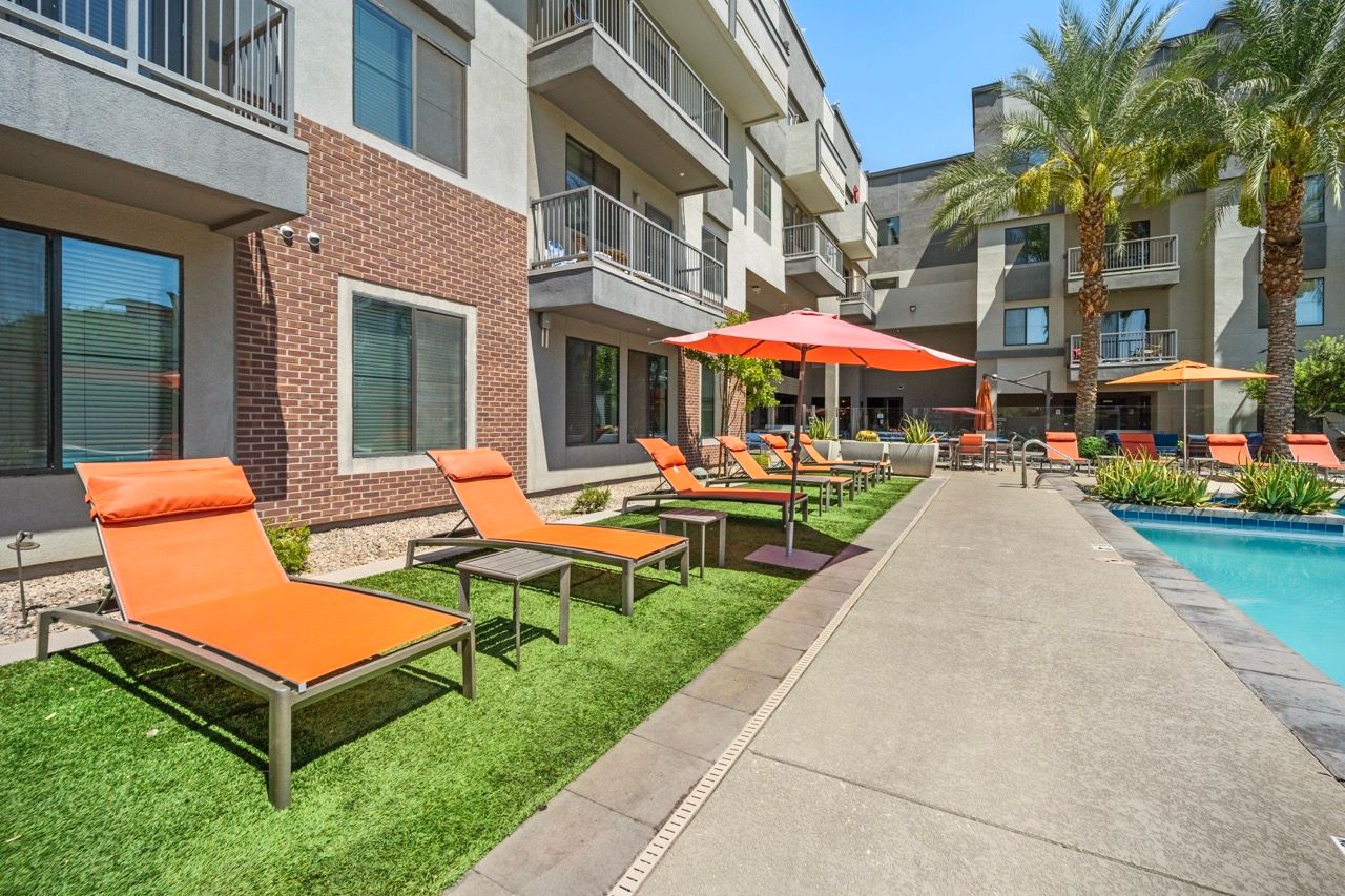 Poolside lounge chairs with orange cushions and umbrellas beside an apartment building.