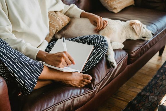 A person in a cream sweater and patterned pants sits on a leather couch, writing in a notebook while petting a sleeping dog.