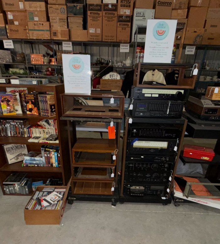 A wooden bookshelf next to an empty media stand and a tall rack of vintage audio equipment in a cluttered storage room.