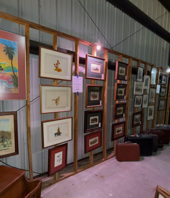 Framed artworks hang on a wooden display wall inside a rustic building, with several vintage suitcases placed on the floor.