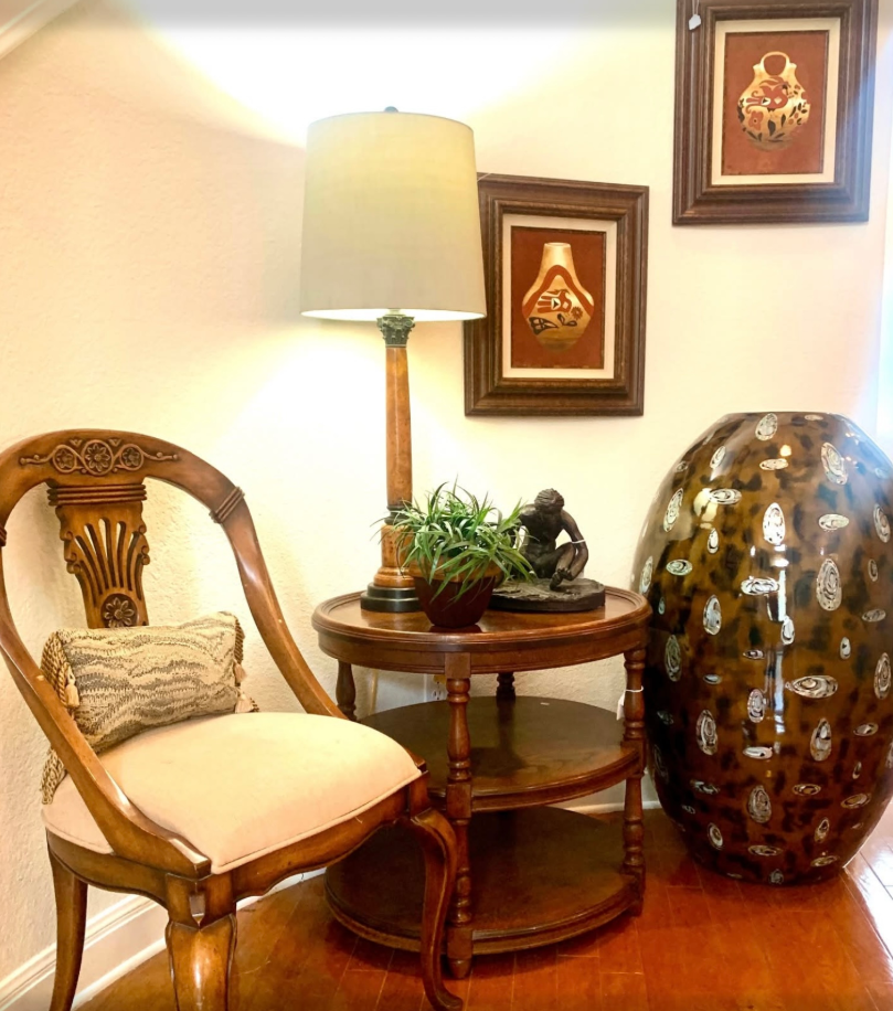 A wooden chair, a two-tiered side table with a lamp and decor, and a large decorative patterned vase in a corner setting.