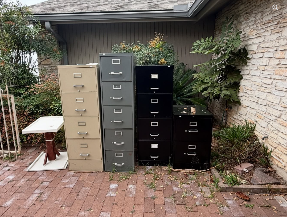 Four filing cabinets of varying heights and colors—tan, gray, and black—stand on a brick patio beside a stone wall.