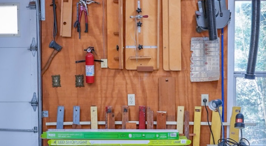 A workshop wall featuring hanging wooden jigs, a fire extinguisher, various hand tools, and colorful clamps on a rack.