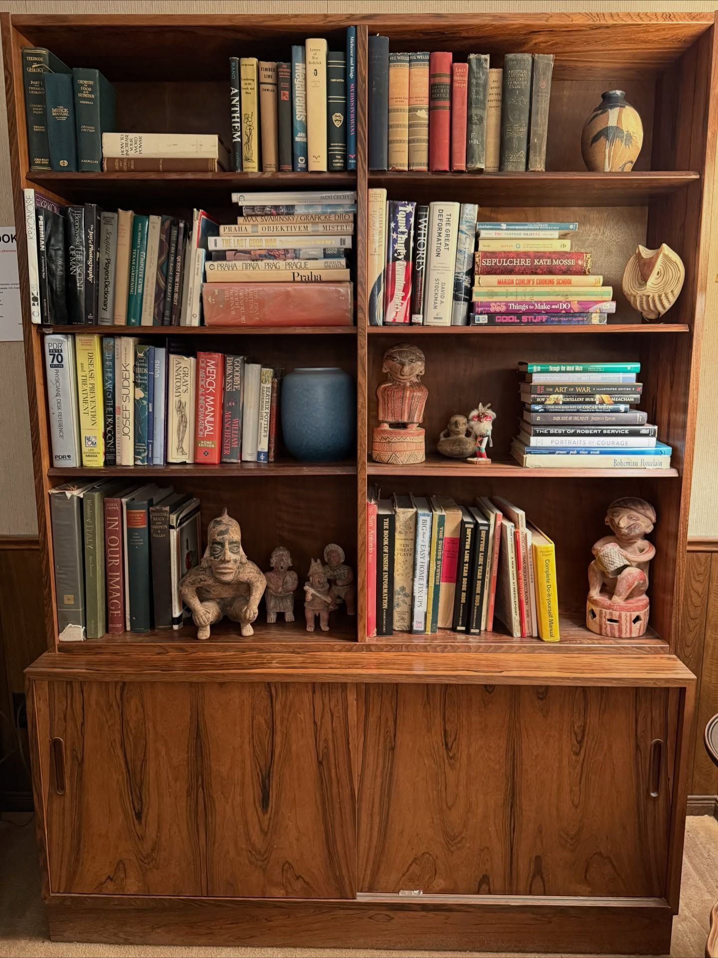 A tall wooden bookcase with open shelving filled with books, small sculptures, and pottery, featuring a cabinet base.