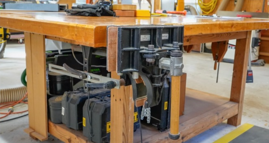 A workbench in a woodshop featuring a mounted black metal vise and various tool storage cases on the lower shelf.