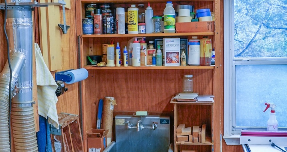 A woodworking workshop featuring a metal utility sink, wooden shelving stocked with various supplies, and a shop vacuum.