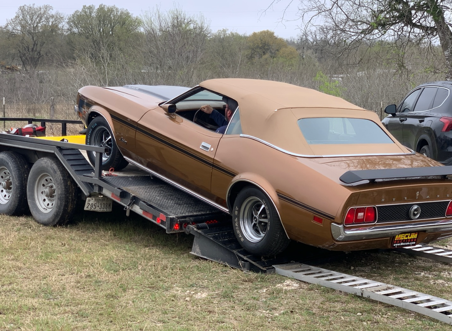 A metallic brown Ford Mustang convertible being loaded onto a flatbed trailer in a grassy, rural setting.