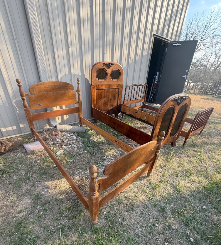 Three wooden bed frames of varying styles are arranged outdoors on a grassy area in front of a metal building.