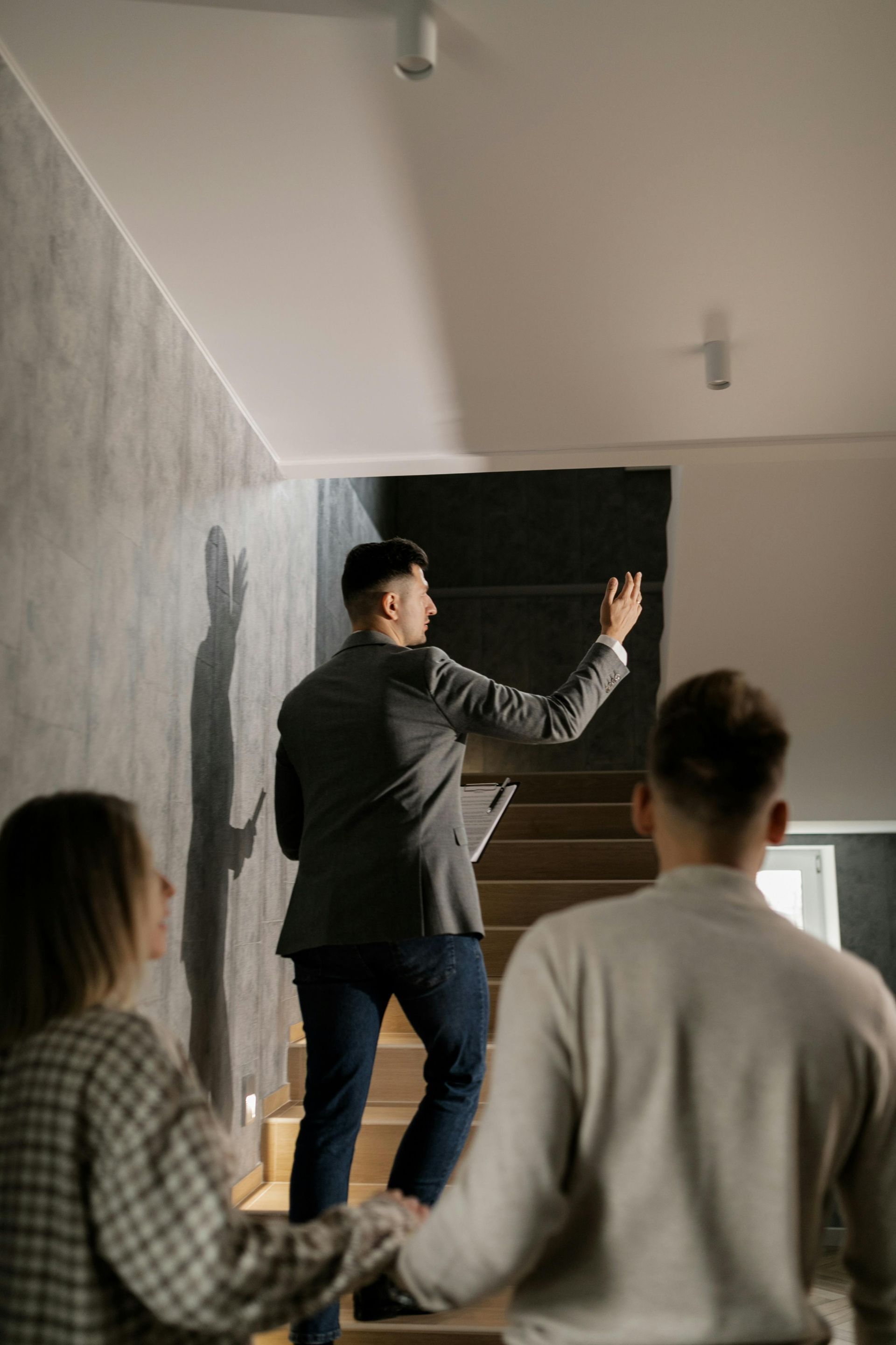 Real estate agent showing a couple a staircase, pointing to the ceiling. Gray wall, wooden steps, and modern lights are visible.