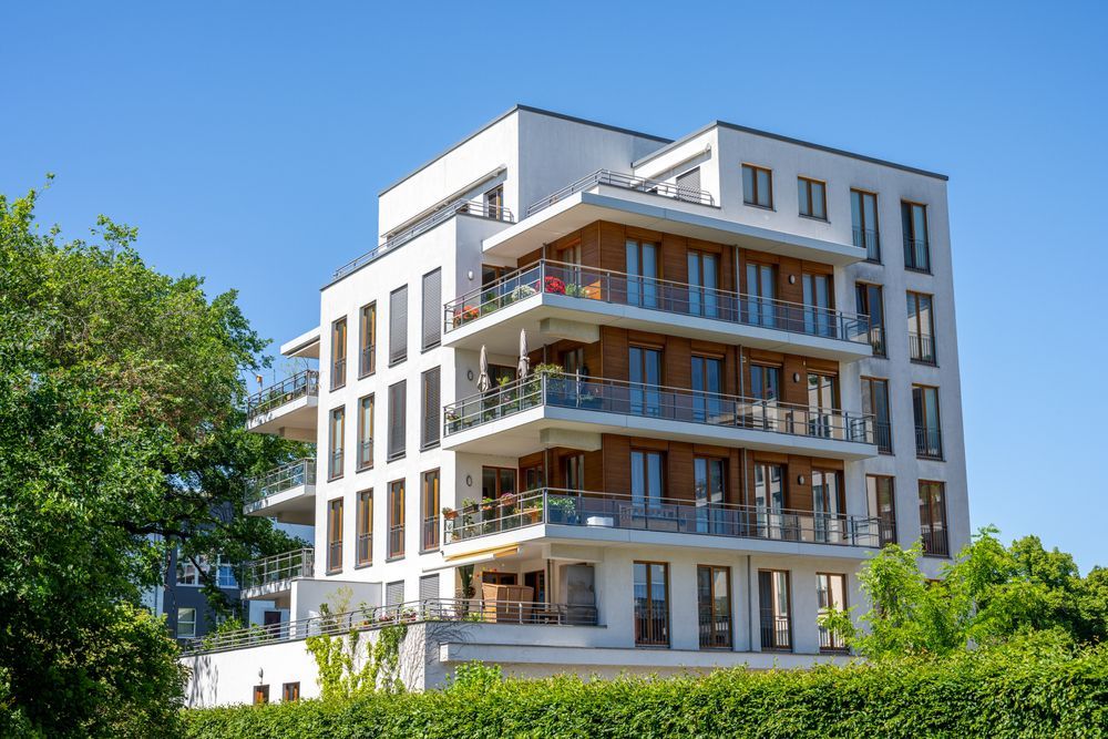 White multi-story apartment building with balconies, set against a blue sky, partially obscured by green trees.