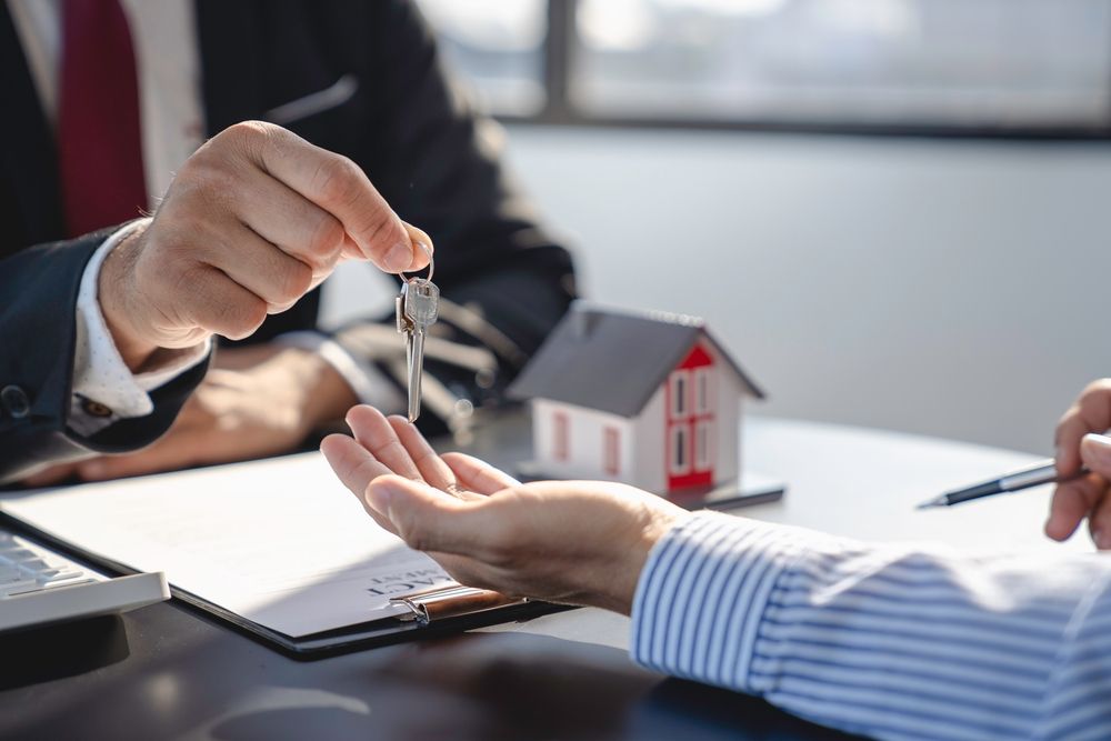 Person handing keys to another person, a model house sits nearby on a desk.
