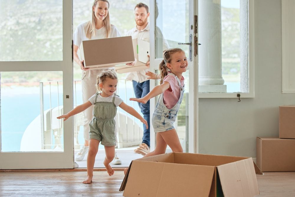 Family moving into a new home; two young girls run excitedly ahead, parents follow with boxes.