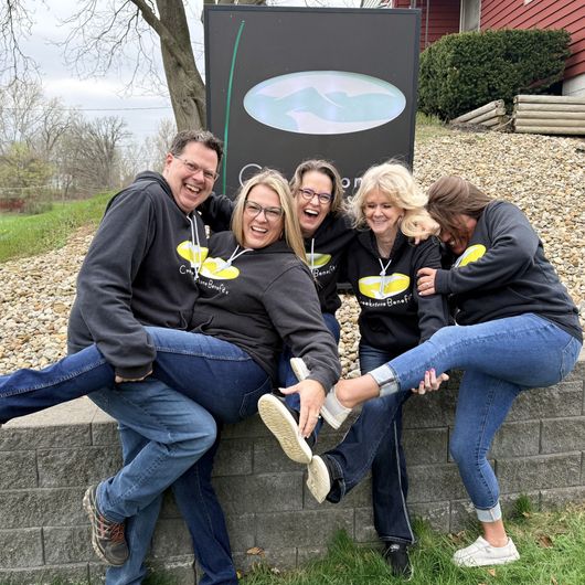Five smiling people in matching black hoodies posing together outdoors by a sign on a stone wall.
