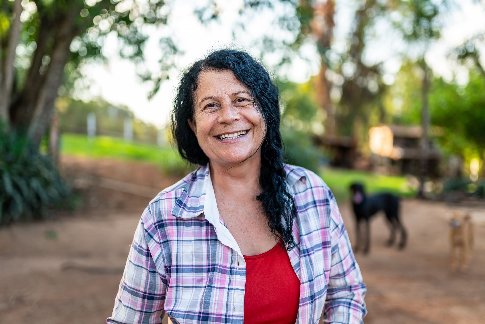 Smiling woman in a plaid shirt standing outdoors on a dirt path with trees and a dog in the background