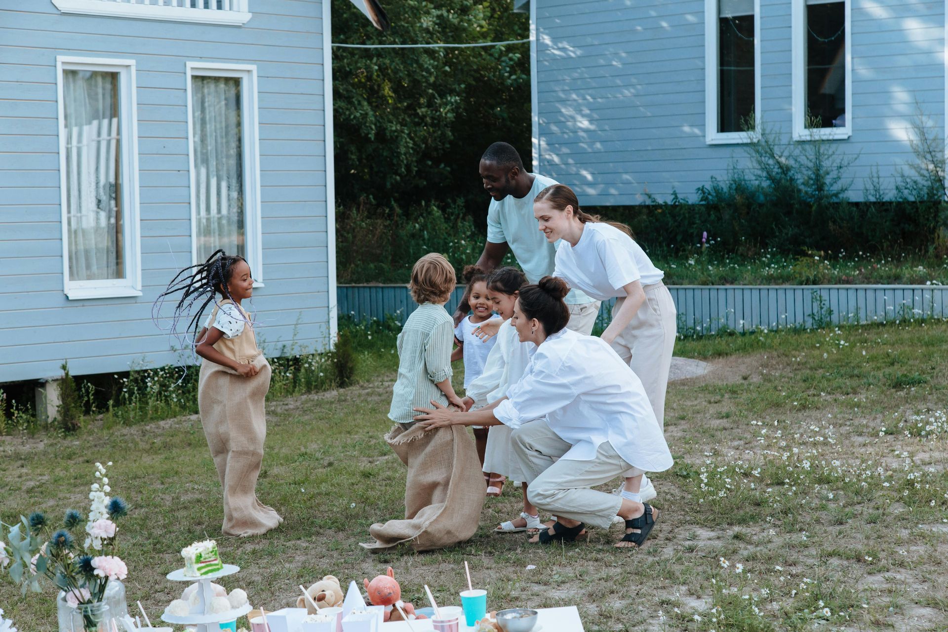 People kneeling with a child in a yard beside a blue house, playing with small items on the ground