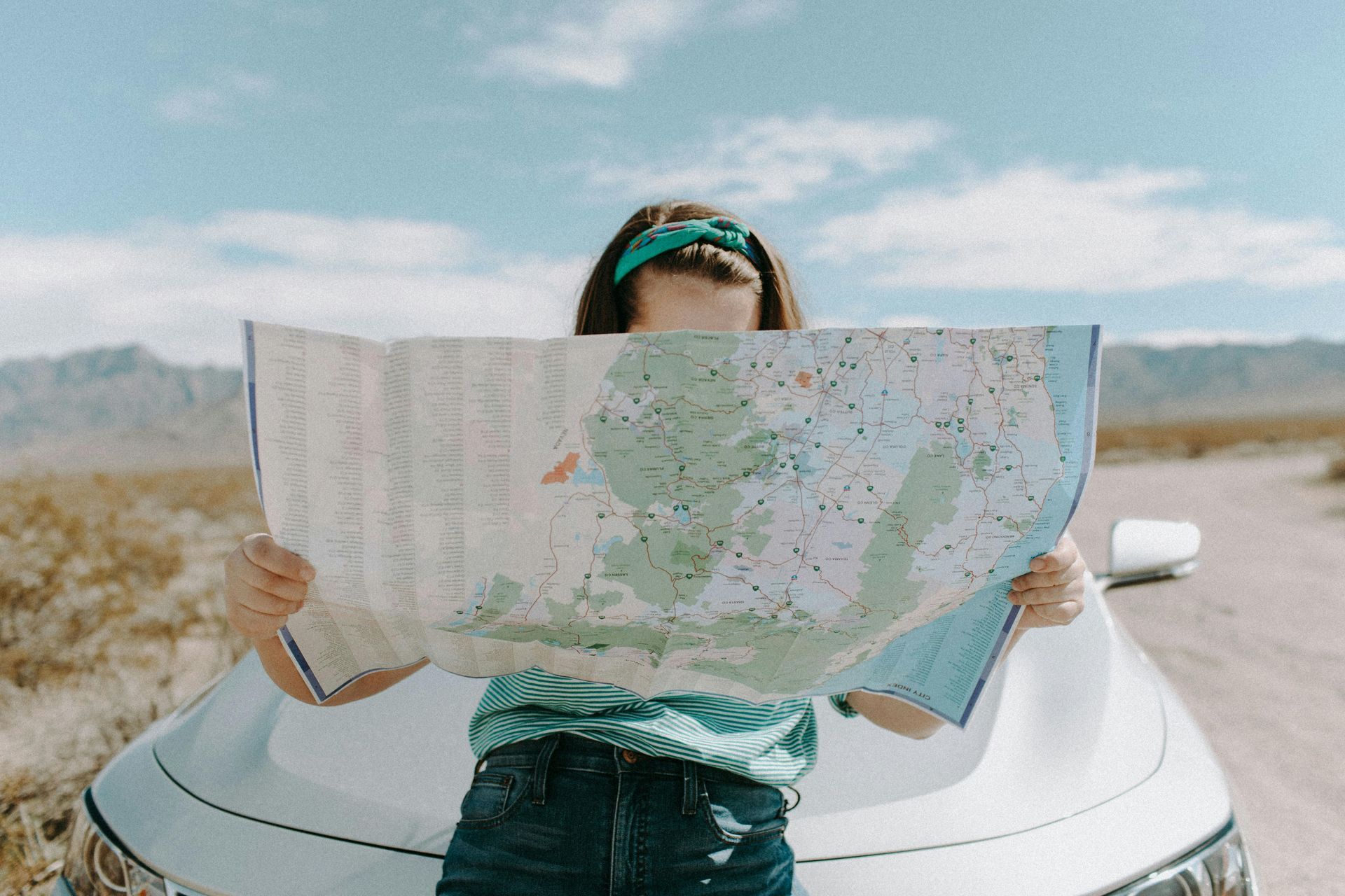 Woman with a bandana over her eyes holds a map over a car in a desert setting.