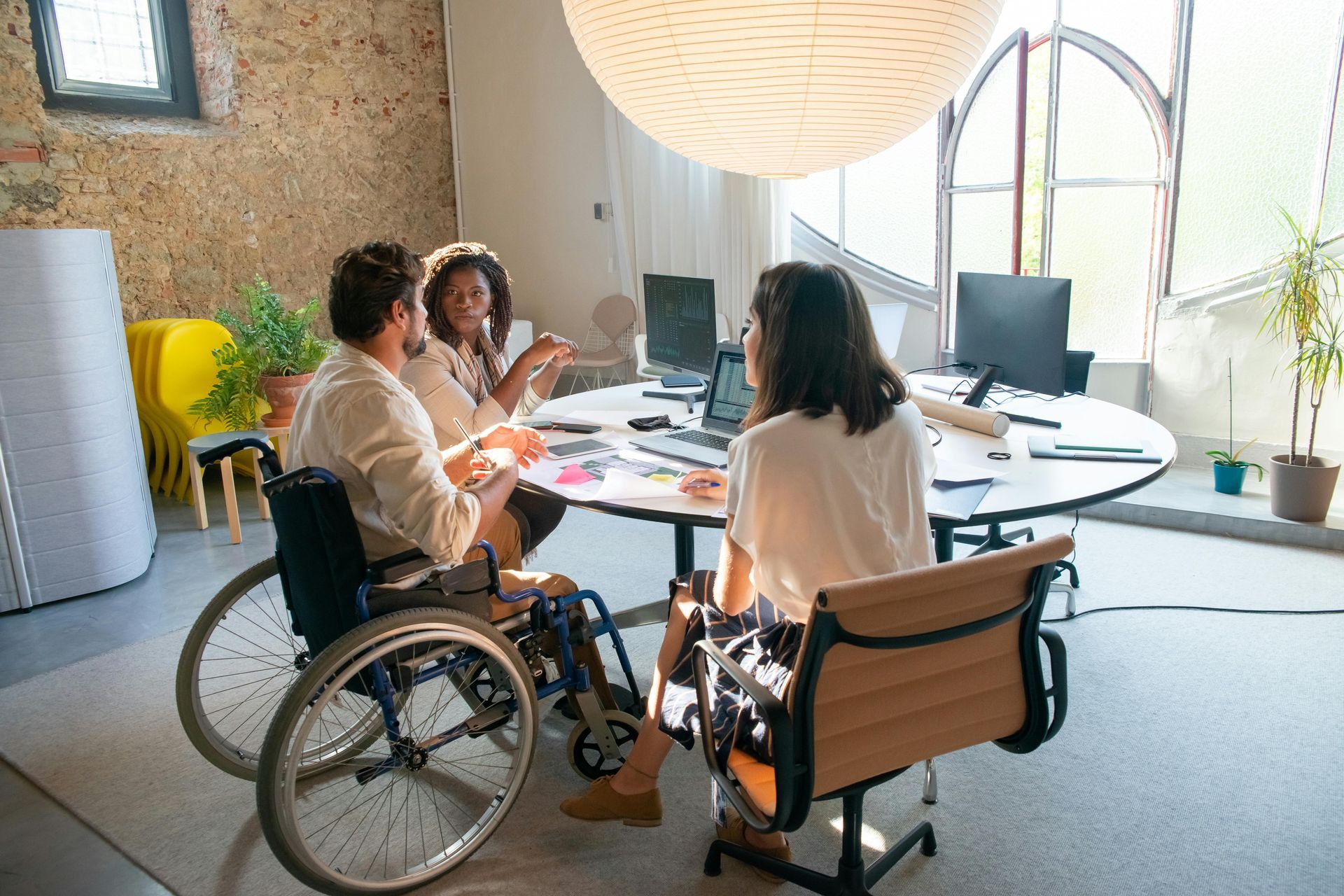 Three people meeting around a table in a bright office, two in wheelchairs, discussing work on laptops.