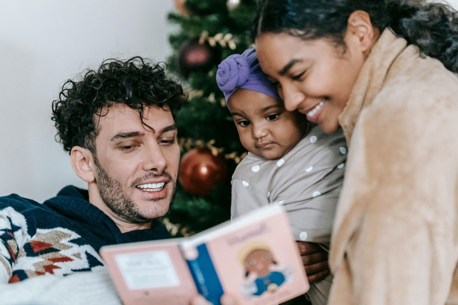 Three people smile while reading a book together near a decorated Christmas tree.