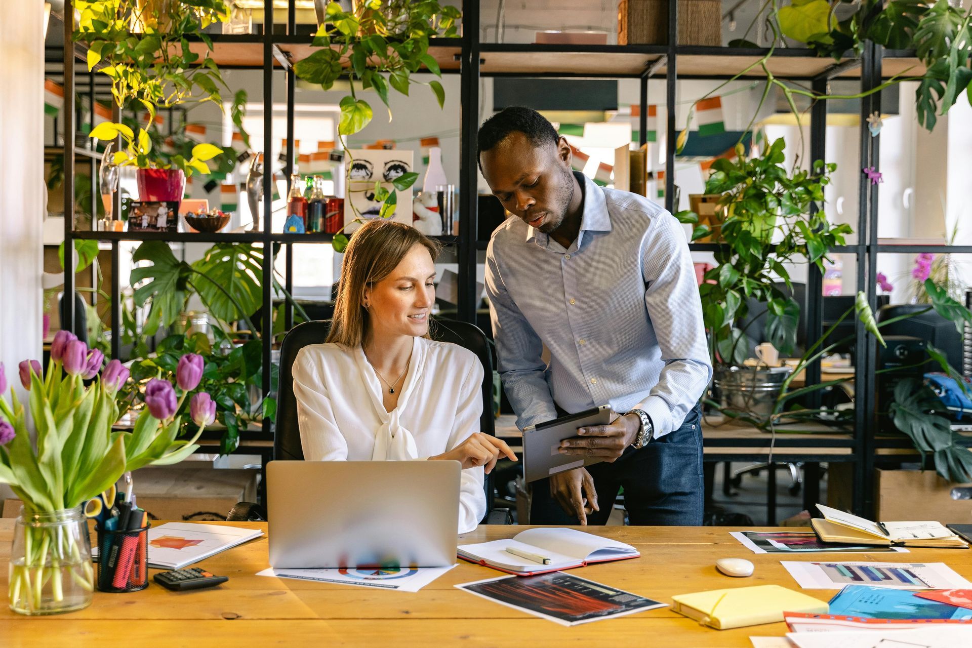 Two coworkers collaborating at a desk in a bright plant-filled office, with laptop and documents.