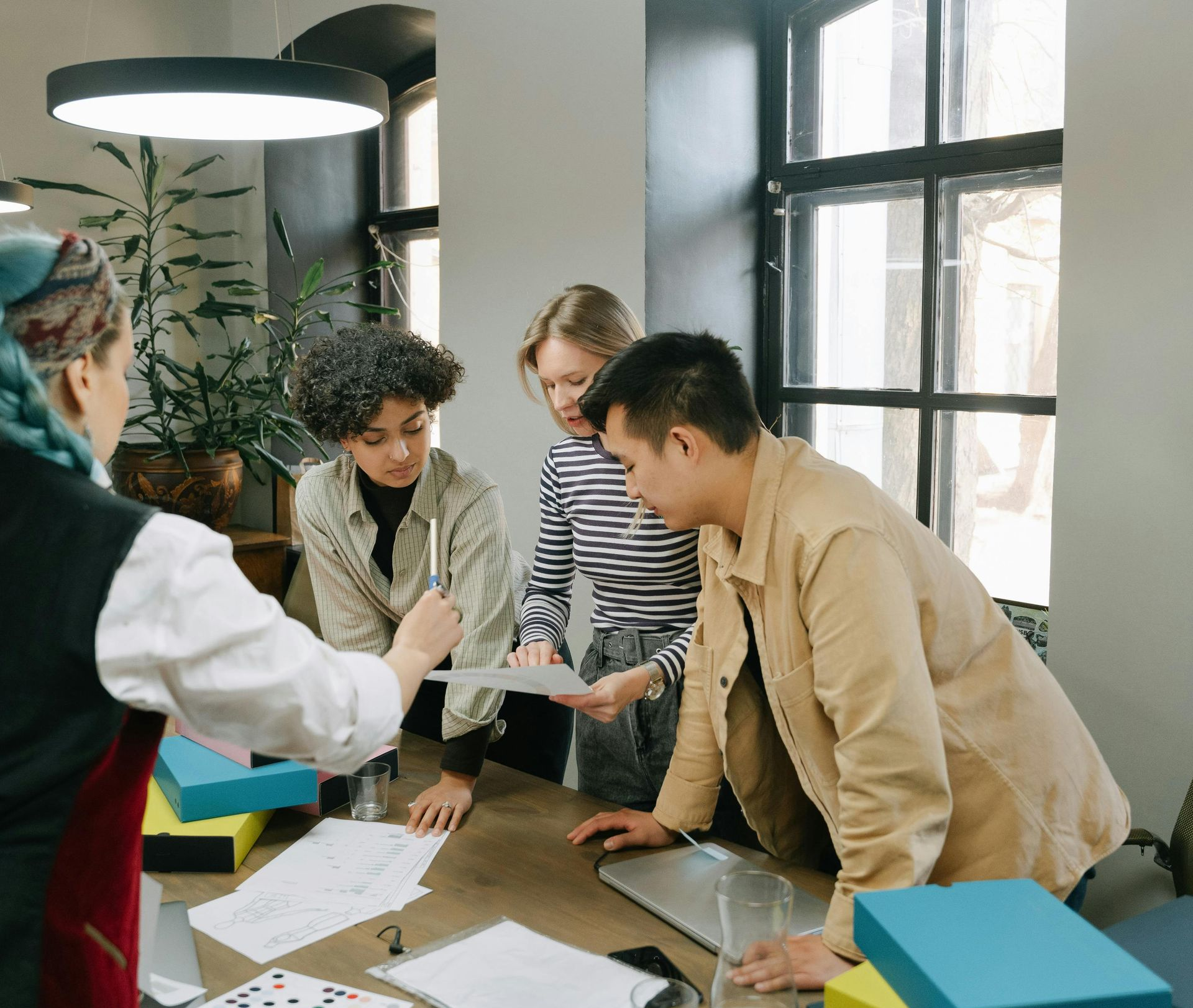 Four coworkers leaning over a table, reviewing papers in a bright office workspace.