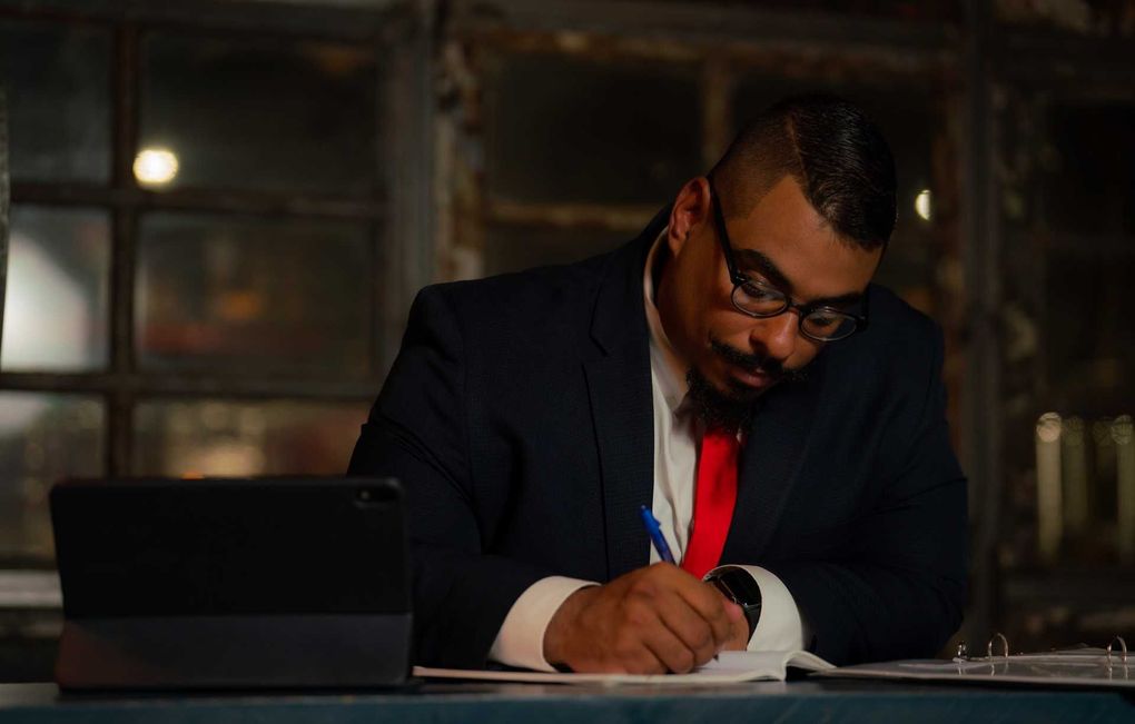 Man in suit, glasses, red tie, writing at a desk, with tablet, near window.