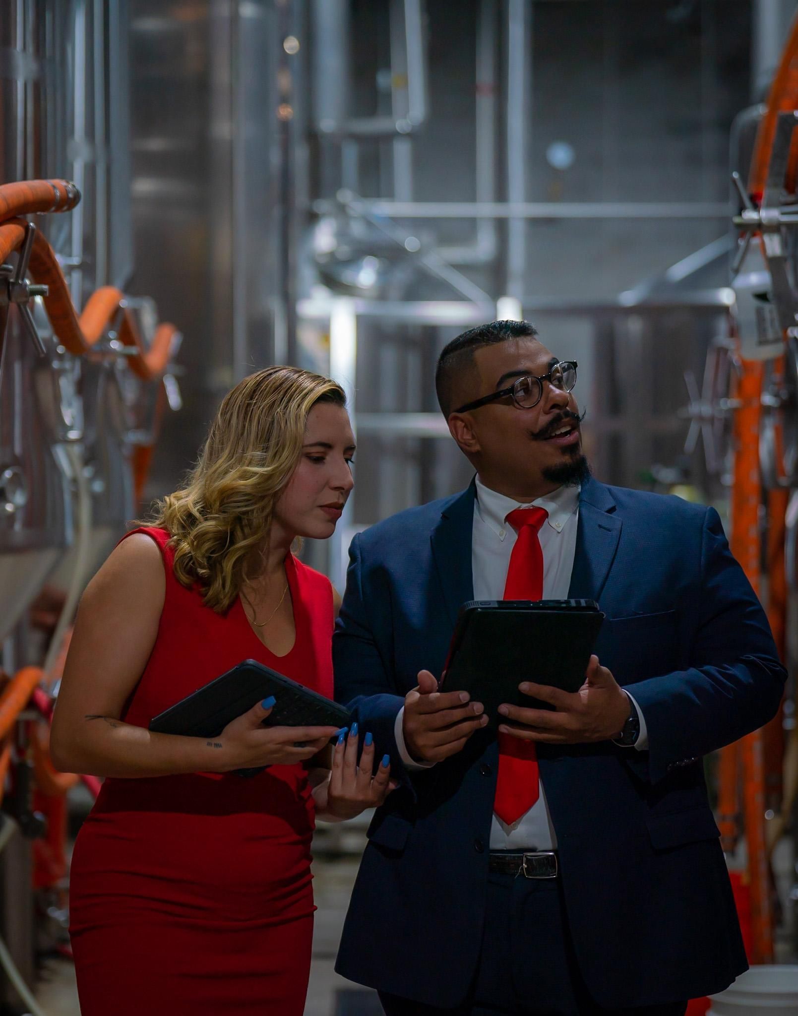 Man in a suit and woman in red dress reviewing documents inside a brewery, looking at equipment.