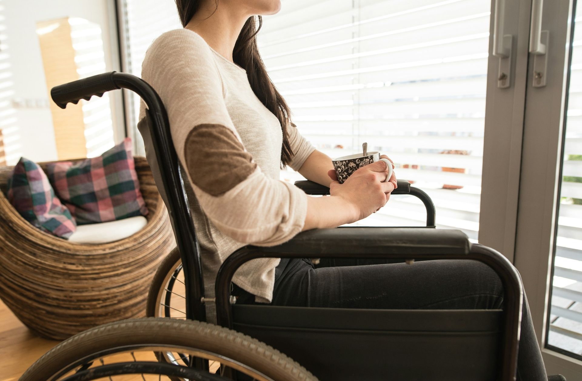 Woman in a wheelchair looking out a window, holding a mug.