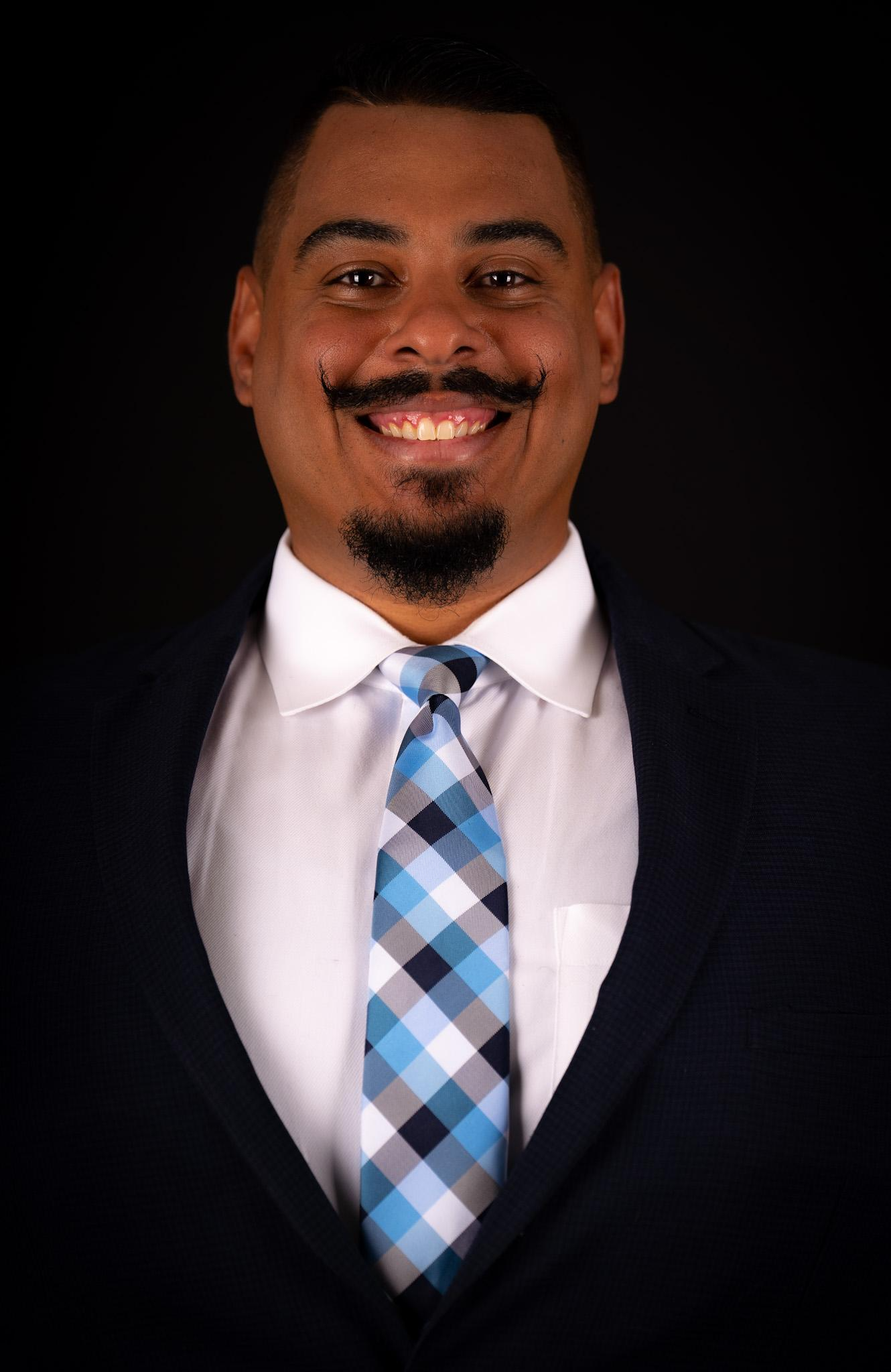 Man with a mustache smiling, wearing a suit and patterned blue tie, against a black backdrop.