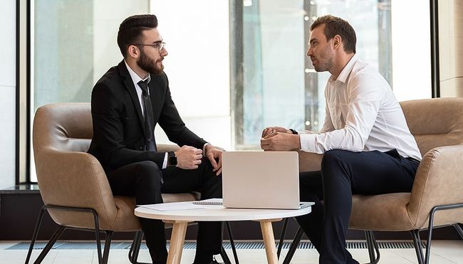 Two men are sitting on a couch talking to each other.