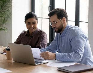 A man and a woman are sitting at a table looking at a laptop.