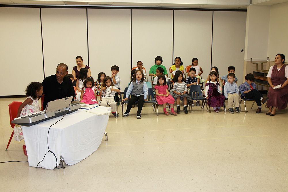 A group of children are sitting in a room with a man playing a keyboard.
