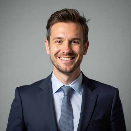 Man in a navy suit and light blue tie, smiling, against a gray backdrop.