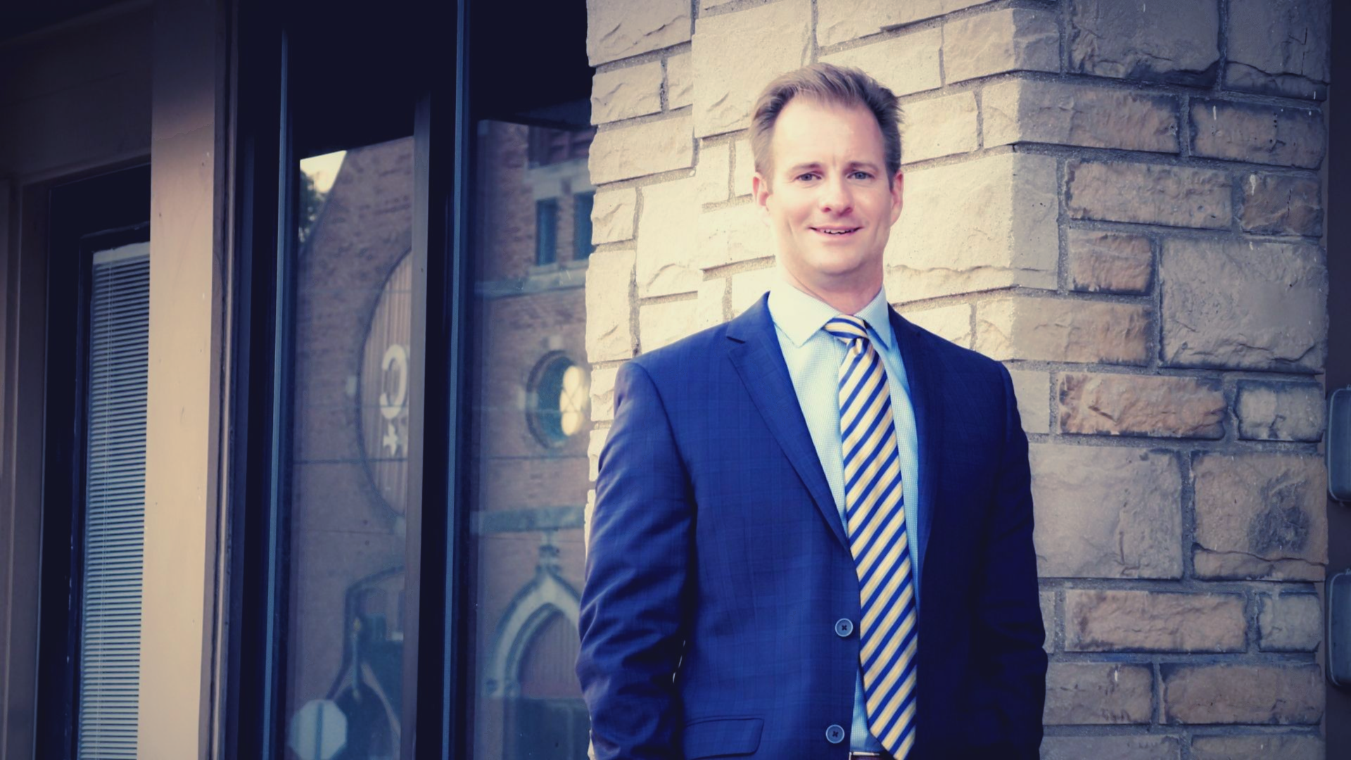 Man in a blue suit and tie, smiling, standing outside a brick building.