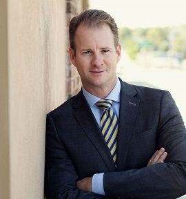 Man in a dark suit with striped tie, arms crossed, standing by a wall in a bright outdoor setting