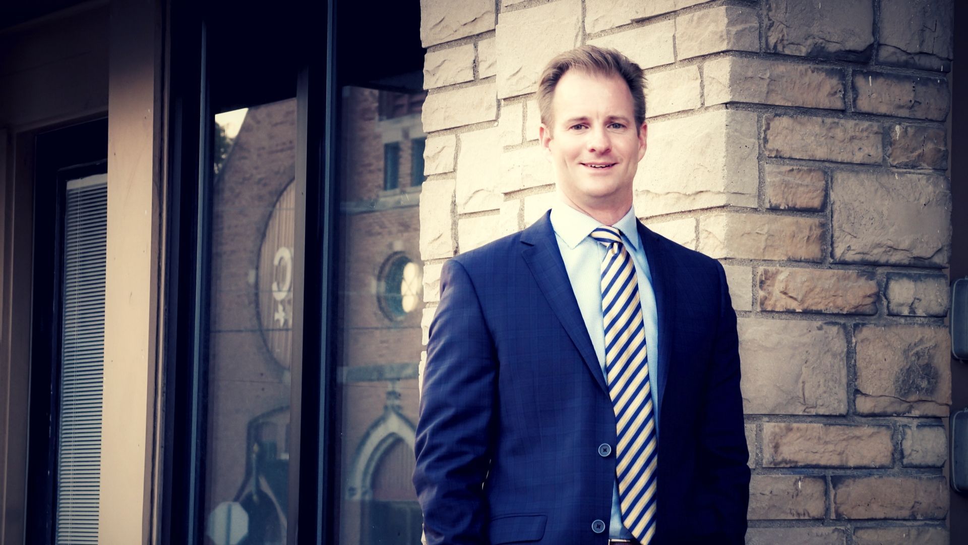 Man in a blue suit and tie, smiling, standing outside a brick building.