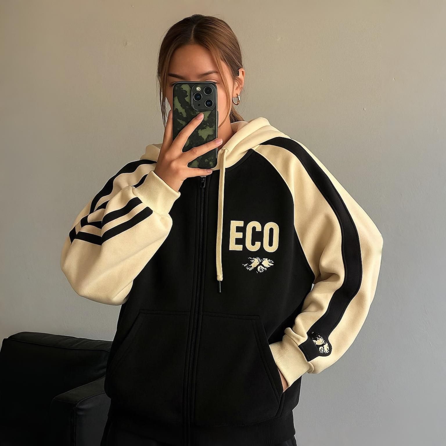 Mujer con sudadera con cremallera de color negro y crema, sosteniendo un teléfono, en un entorno interior.