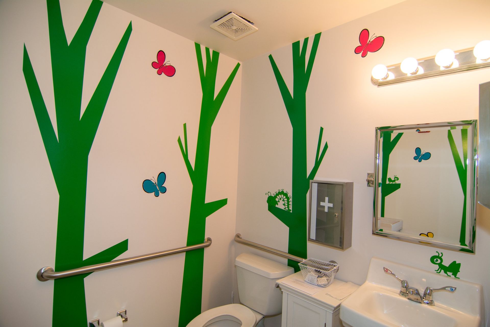 Photo inside the comfort room. white walls with green trees and butterfly stickers, white toilet bowl, sink, and  amirror 