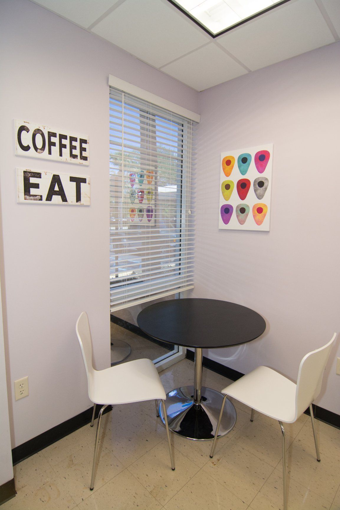 Round table with two white chairs in the corner beside a glass window with blindes. on the right wall hands a painting of an av