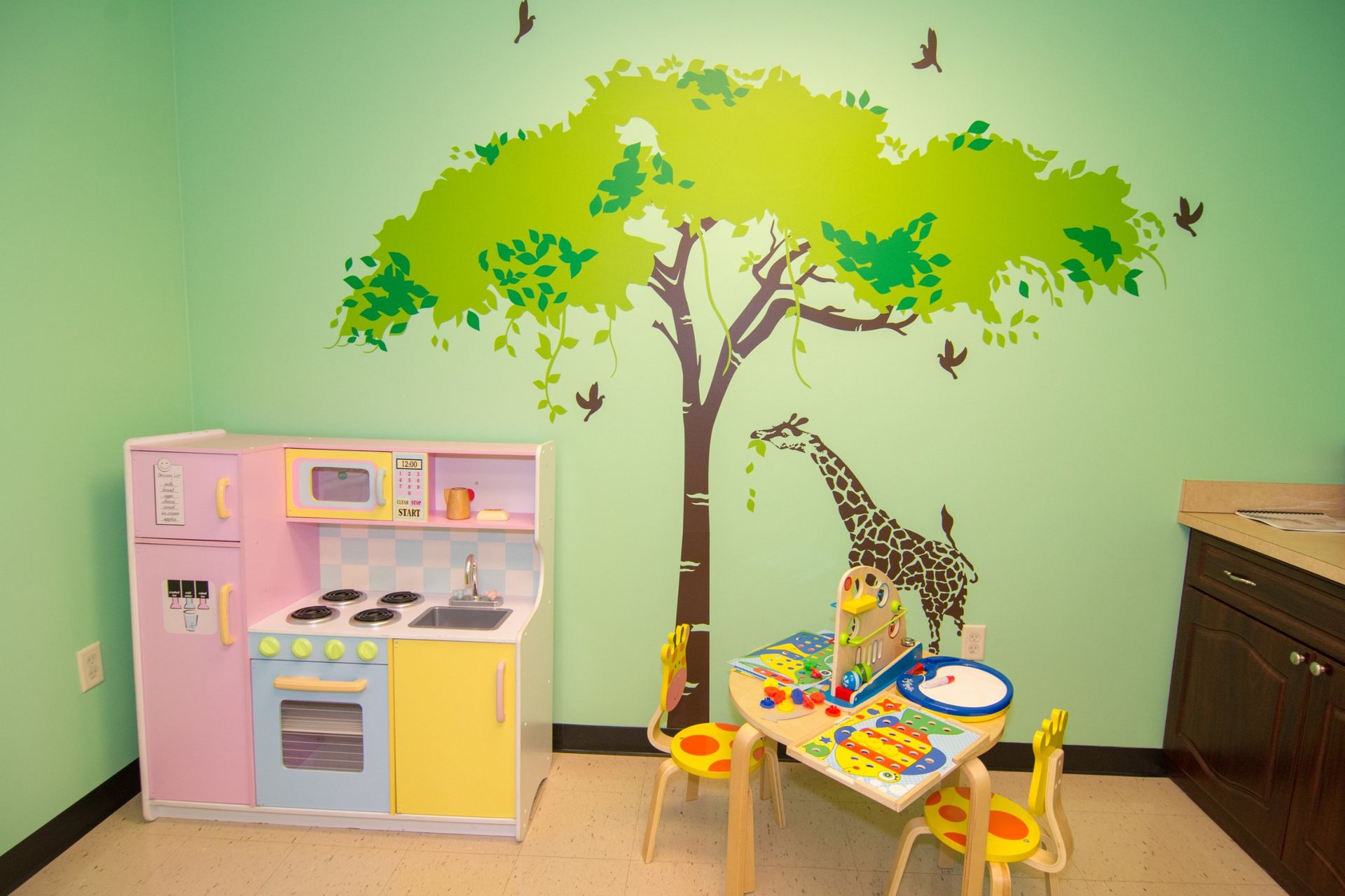 kitchen play set against a light green wall with a tree and giraffe sticker. beside a table with two chairs and sink on the further right.