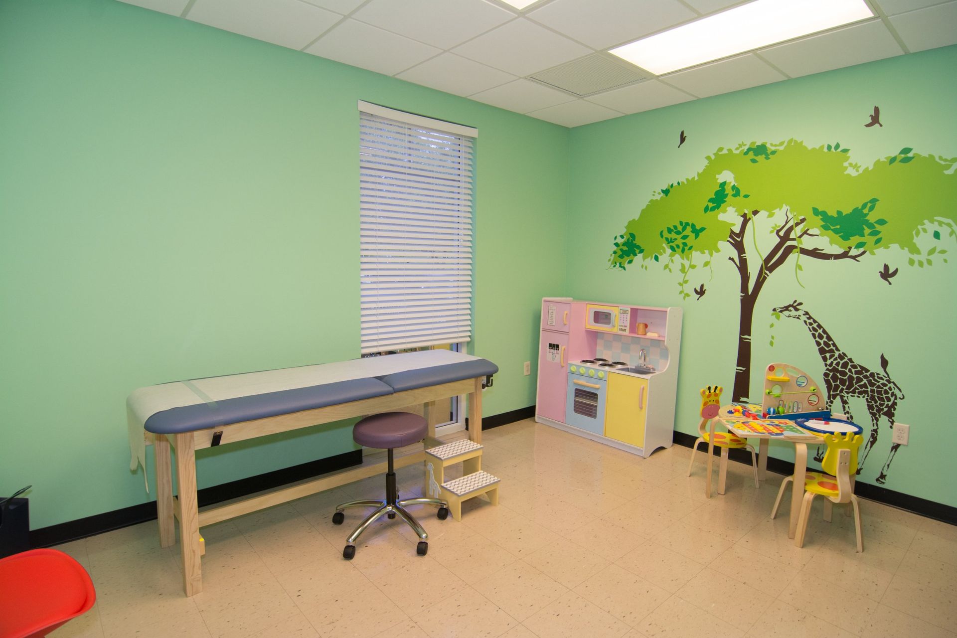 Photo of the consultation room with light green walls with a tree and giraffe sticker, showing the examination bed on the left against the wall beside the window, one the other corner of the room is the play kitchen set, table and chairs for kids 