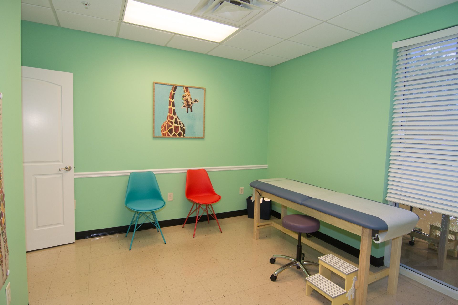 photo of the consultation room. red and blue chairs below a painting of a zebra hanging from the light green wall beside the white door. the bed is on the right side beside the window
