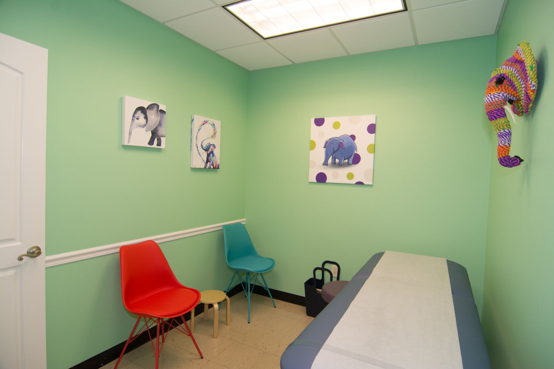 photo of the consultation room showing red and blue chairs against the wall, a bed on the opposite directio, three drawings of elephants handing on the wall and one elephant head sculpture. 