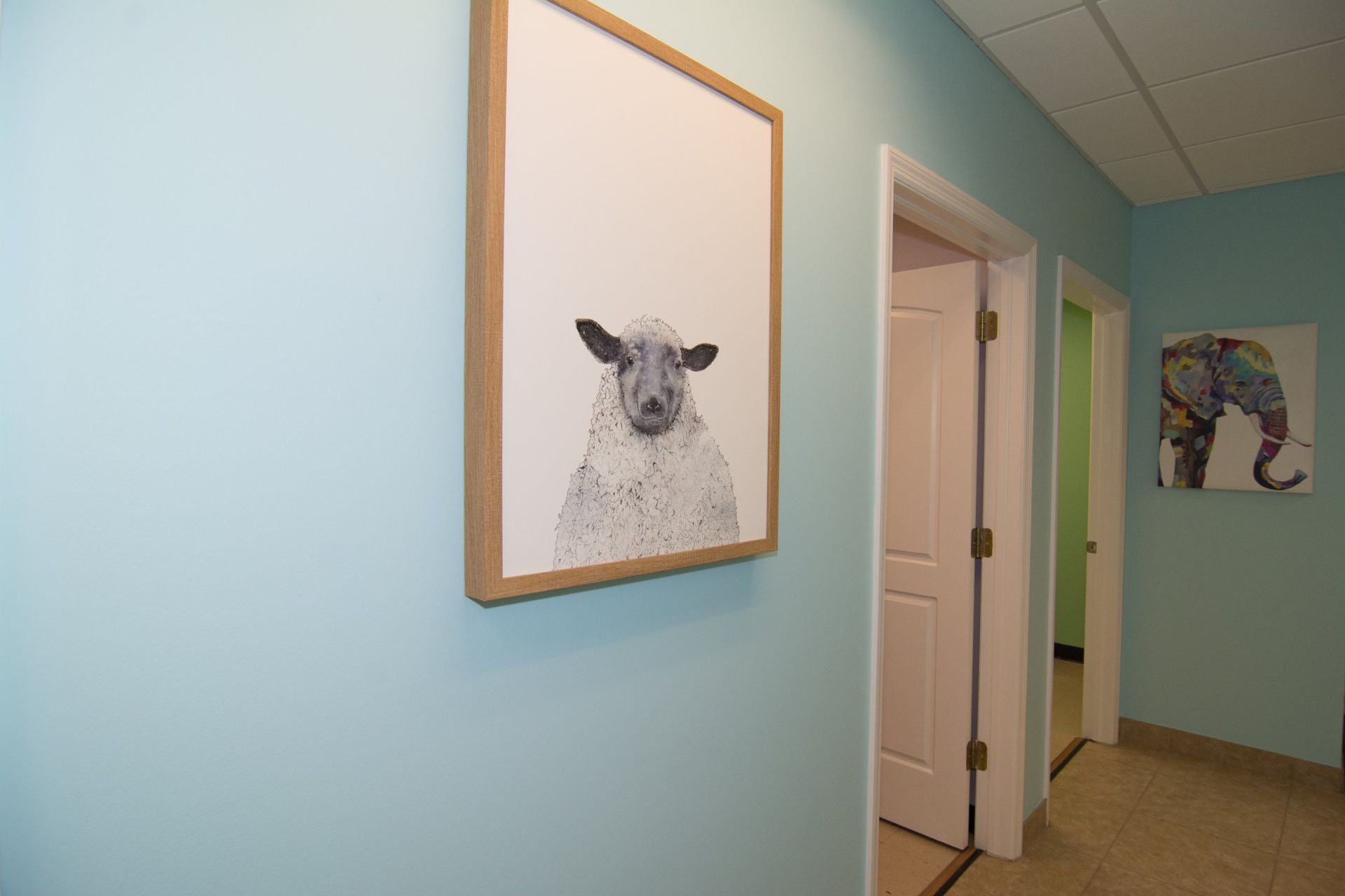Photo of the lobby, light blue walls, pencil drawing of a sheep handing on the wall,  two doors on the farther left, and painting on an elephant on the wall across the hall